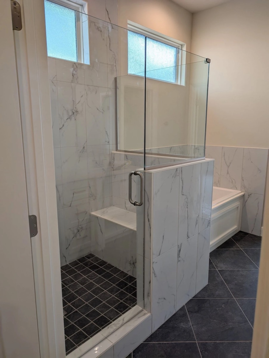 Glass shower door and fixed panel on a marble-look knee wall with a brushed metal handle and a built-in bench, installed by Bob's Shower Plus