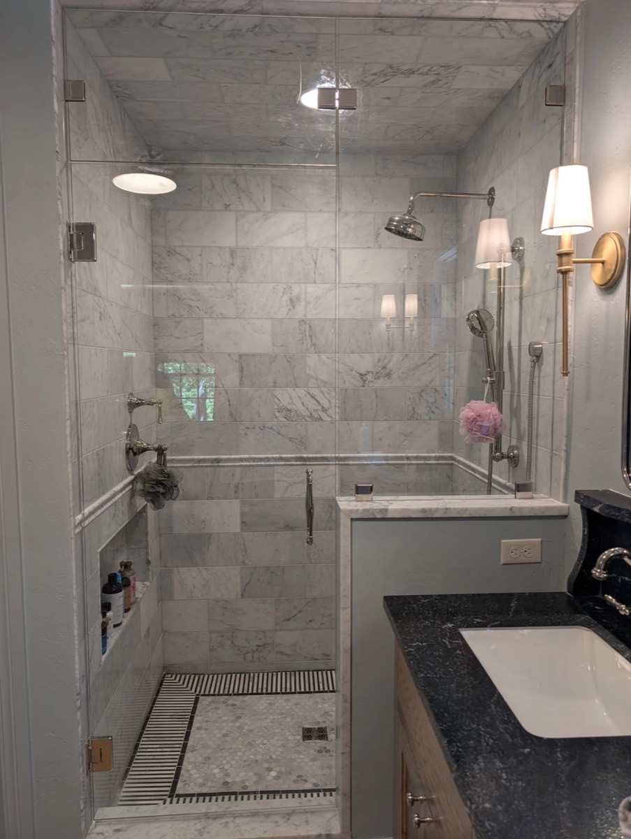Frameless glass shower door with a glass transom panel to the ceiling and polished hardware in a marble shower, installed by Bob's Shower Plus