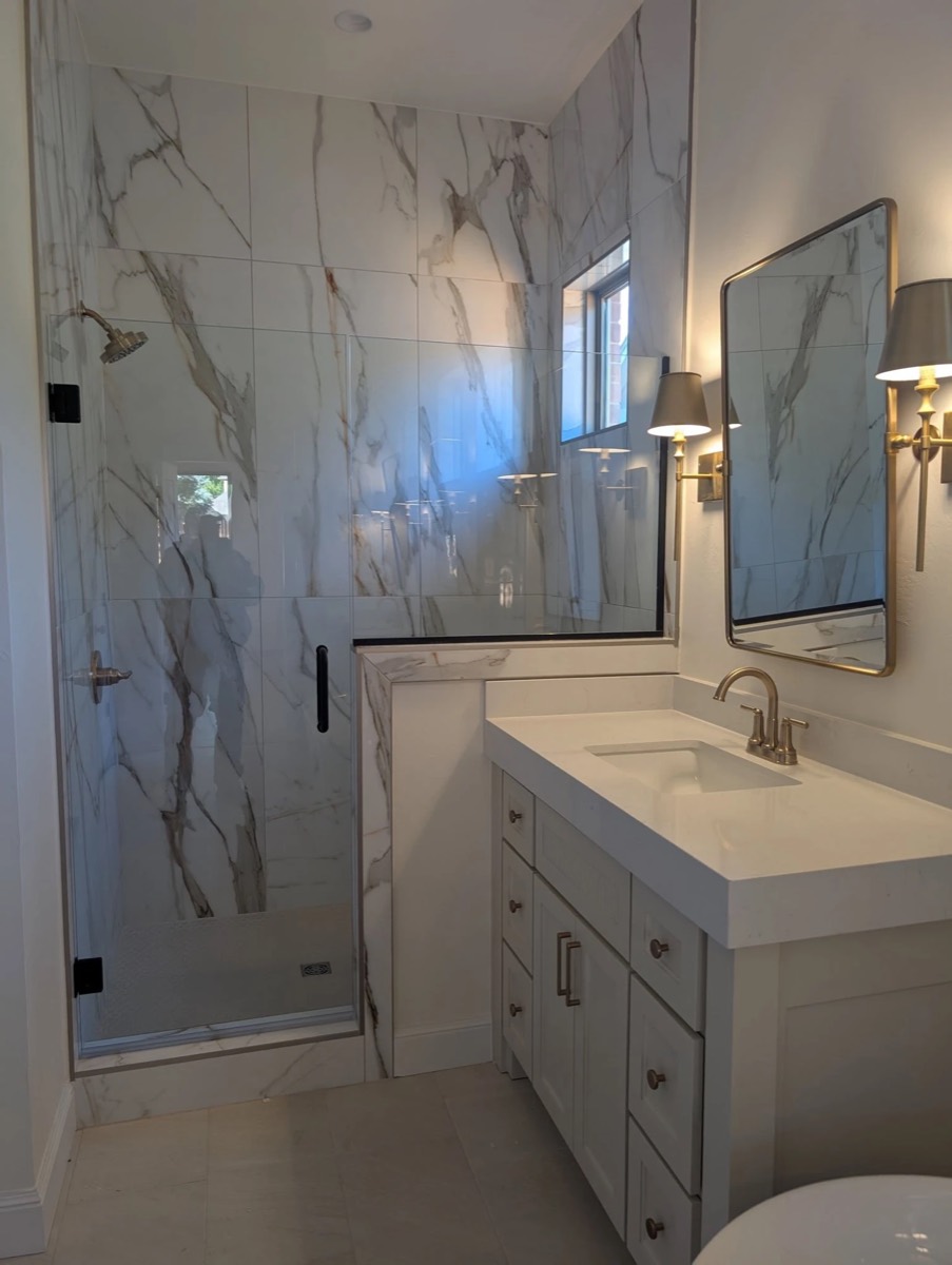 Frameless glass shower door and half panel on a knee wall with black handle and hinges beside a white vanity, installed by Bob's Shower Plus