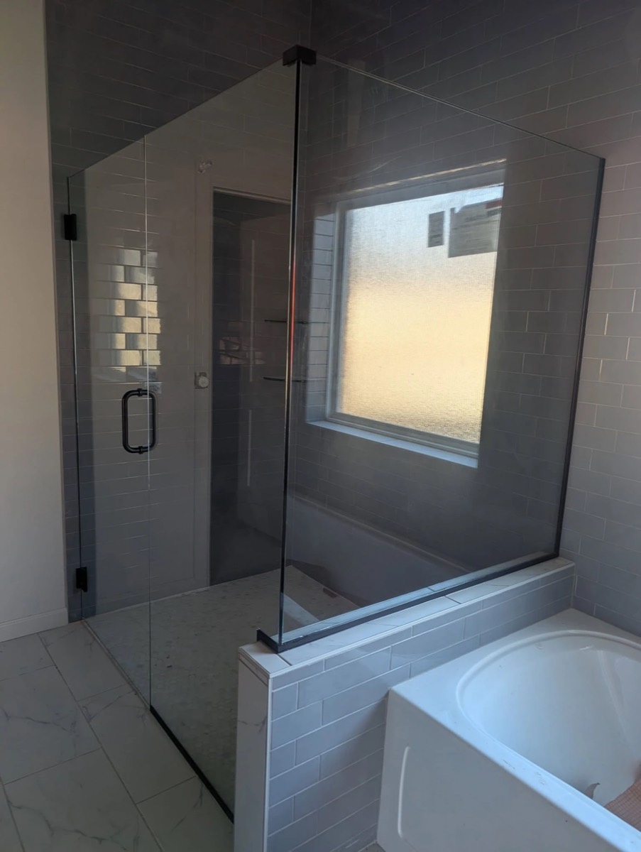 Frameless glass corner shower on a tiled knee wall beside a bathtub, with matte black hinges and handle, installed by Bob's Shower Plus