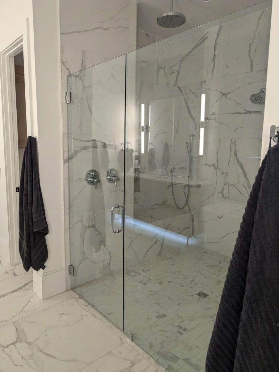 Large frameless glass shower door and fixed panel with chrome hinges and handle in a marble-look walk-in shower, installed by Bob's Shower Plus